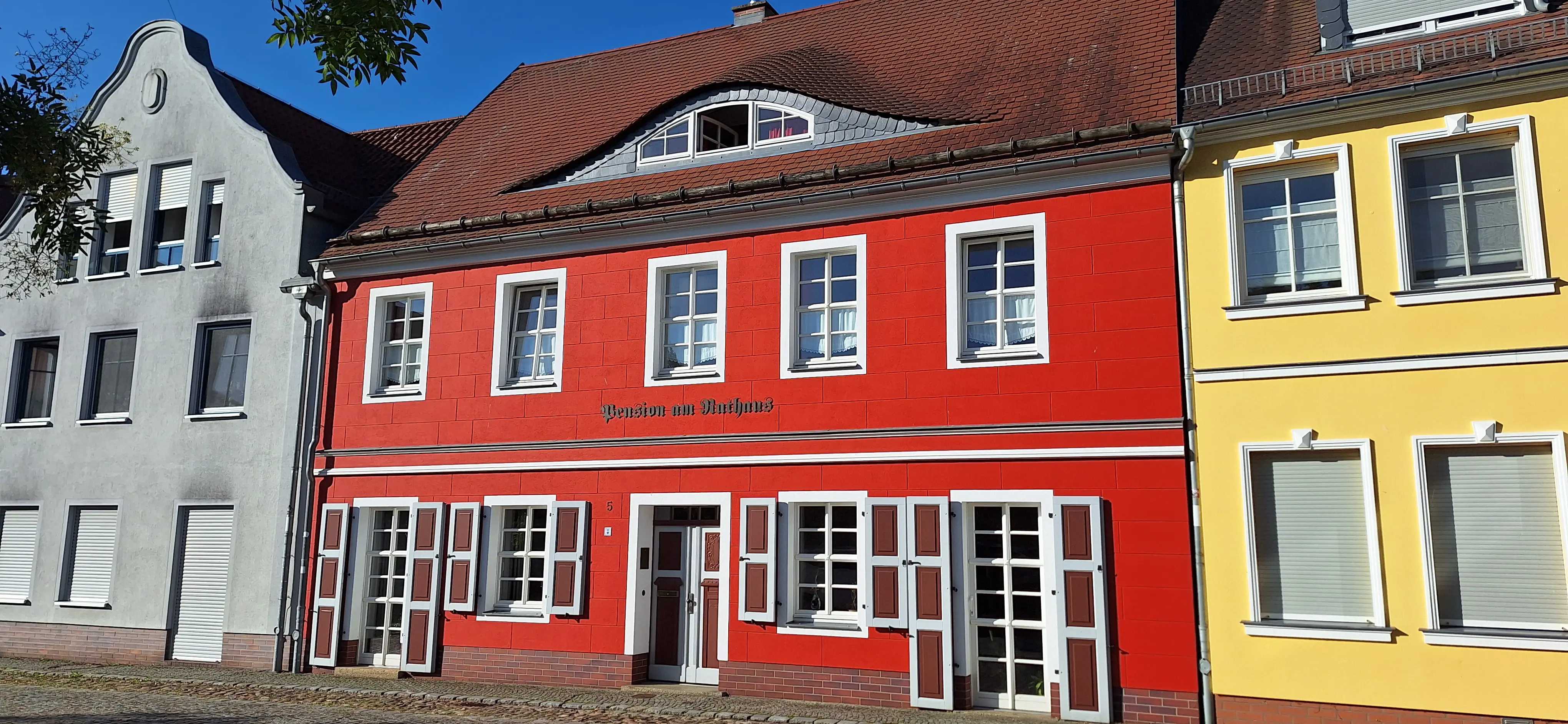 Historisches Gebäude mit roter Fassade, weißen Fensterrahmen, braunen Fensterläden und rotem Ziegeldach in geschlossener Häuserzeile.