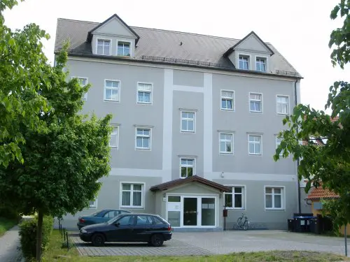 Mehrgeschossiges Wohnhaus mit hellgrauer Fassade, weiß abgesetzten Fenstern und steilem Satteldach, umgeben von Bäumen.